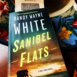 A paperback version of the book Sanibel Flats by Randy Wayne White, with a Doc Dord's Rum Bar & Grille menu as a bookmark, on top of a straw sun hat with a flowery lining.