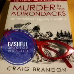 Am image of the book Murder in the Adirondacks by Craig Brandon with a copy of read cat-eye glasses on top of the book cover and a badge reading "Bashful Adventurer Book Club."
