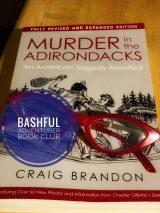 Am image of the book Murder in the Adirondacks by Craig Brandon with a copy of read cat-eye glasses on top of the book cover and a badge reading "Bashful Adventurer Book Club."